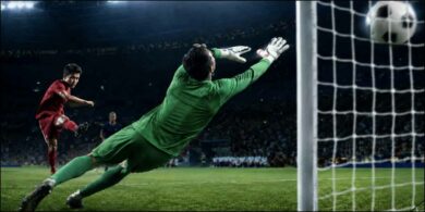 Football player strikes the ball toward goal as the goalkeeper narrowly misses the save.