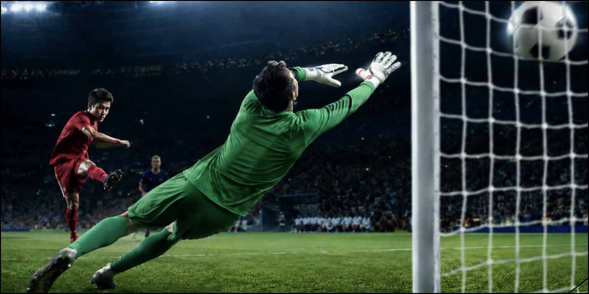 Football player strikes the ball toward goal as the goalkeeper narrowly misses the save.